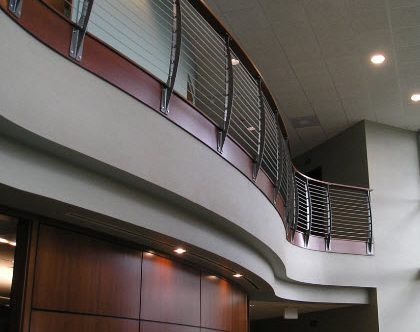 Curved second story loft with the curved Keuka style curvaceous cable railing. The railing is fascia mounted along a curving mezzanine. The Loft has wood cladding that the railing posts are mounted to. Creating a warmth and contrast.