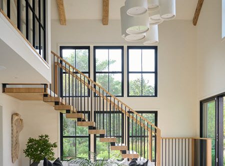 Double stringer floating staircase with spindle railings and white oak treads and handrails in a contemporary setting. The expansive windows allow for natural light to flow throughout the room with white couches.