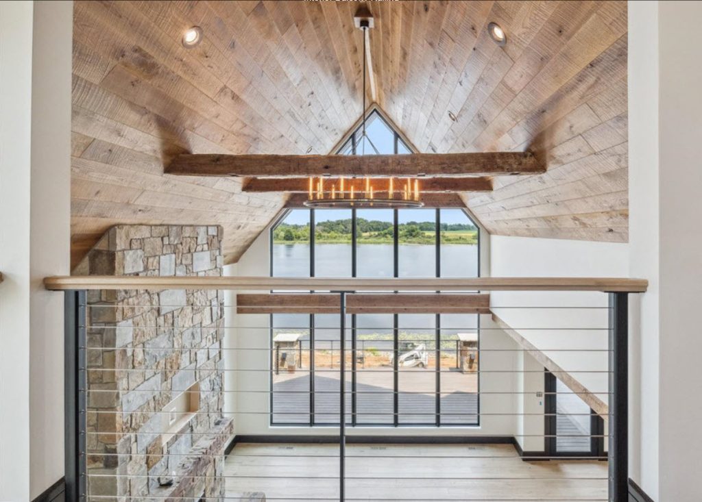 Balcony cable railing overlooking a rustic great room with stone fireplace, wood ceiling and large windows.