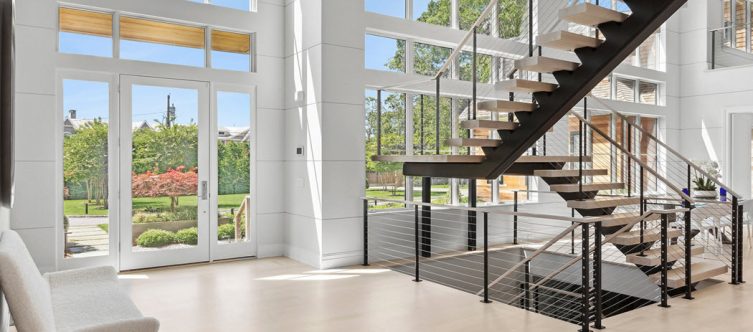 Large great room in a modern home with expansive windows looking ort to a blue sky and landscape. Centrally located in the room is a monostringer staircase that appears to float. The stairs feature a black single stringer and black railing posts with a cable rialing and white oak wood treads that match the floor.