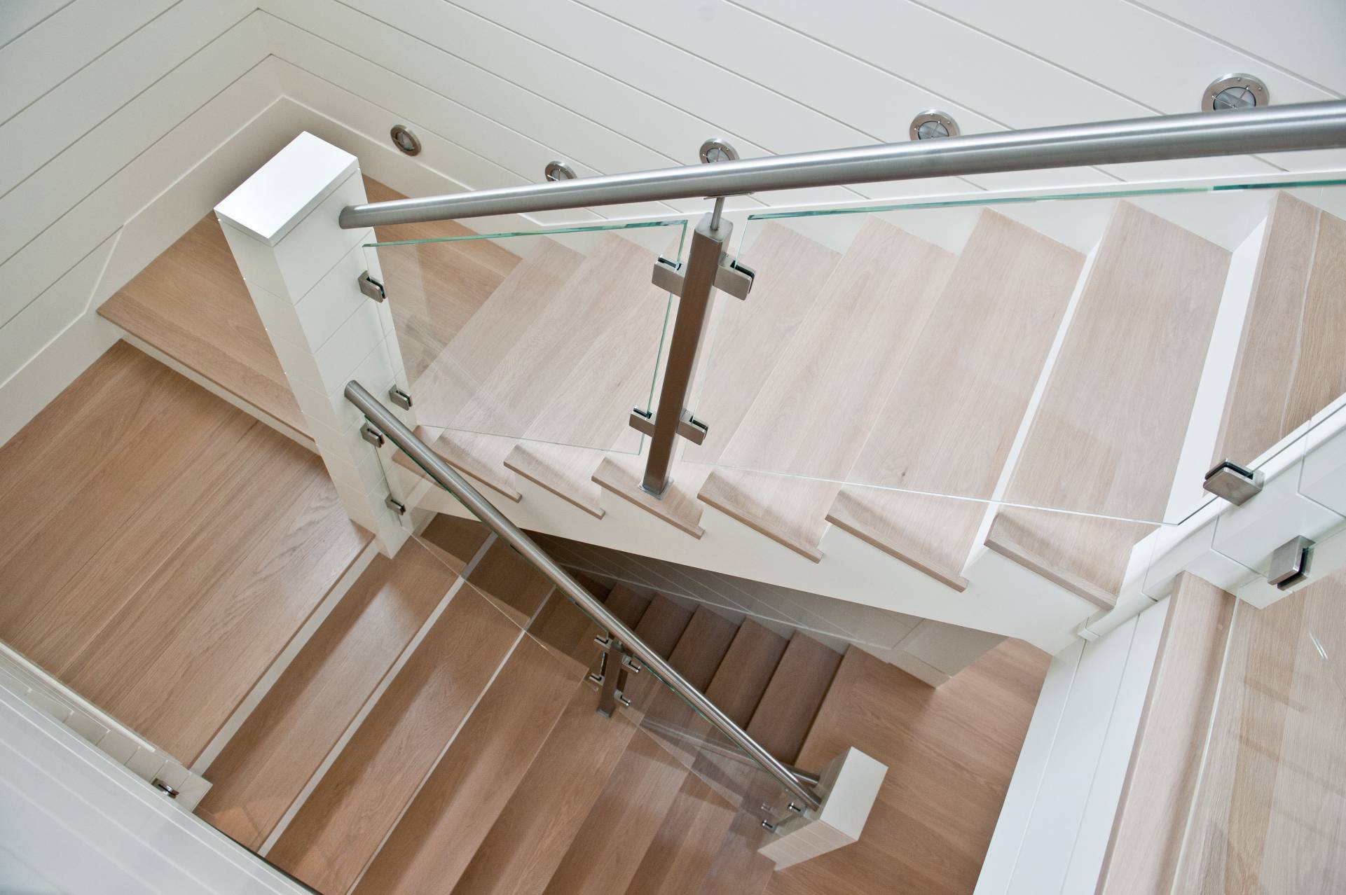 Nautical railing style with round stainless steel handrail and stainless steel glass clamps. The  railing is attached to large wood newel posts complementing a wood floor and stair treads