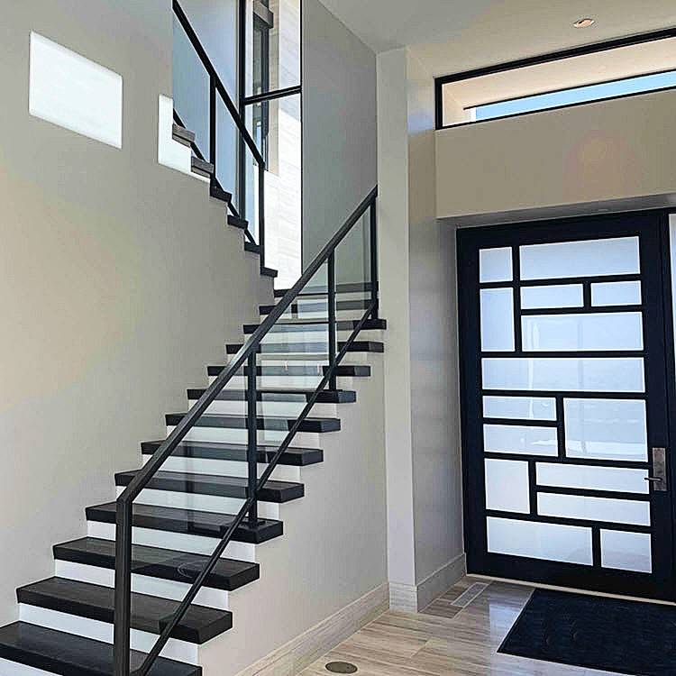 Framed glass railing on dark wood treads with closed risers. The black frame complements the modern door with a glass window and geometric pattern all set in a room with white walls.