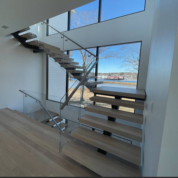 Modern floating single stringer staircase transitions up a window wall with wood treads that match the floor and glass railings 